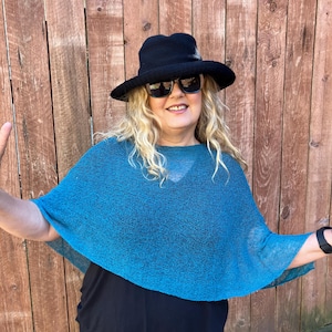 May include: A teal blue knit poncho with a loose, semi-sheer texture, worn over a solid black top. The outfit includes a black wide-brimmed hat and dark-tinted sunglasses, creating a layered look with contrasting colors and varied fabric textures.