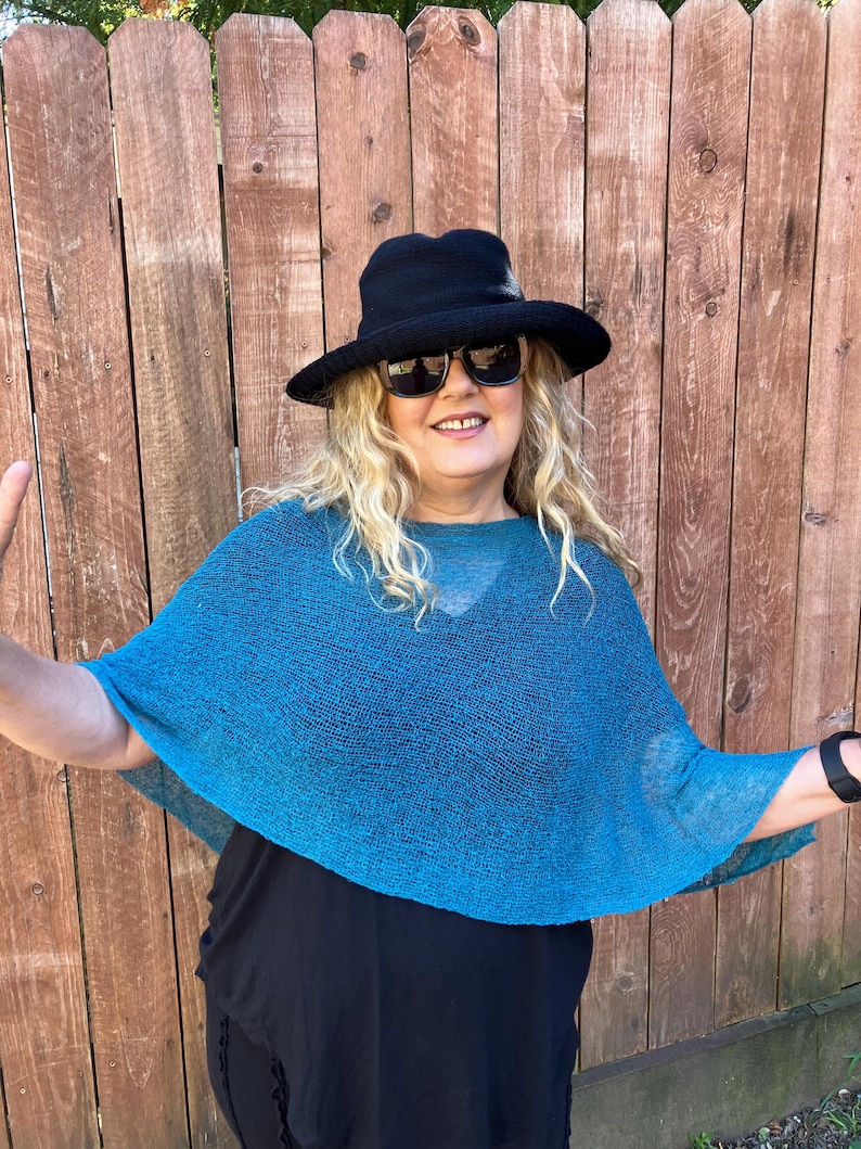May include: A teal blue knit poncho with a loose, semi-sheer texture, worn over a solid black top. The outfit includes a black wide-brimmed hat and dark-tinted sunglasses, creating a layered look with contrasting colors and varied fabric textures.