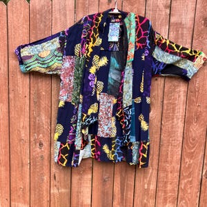 May include: A multi-colored patchwork kimono with a blue background. The kimono has a variety of patterns, including pineapples, animal prints, and floral designs. The kimono is hanging on a wooden fence.
