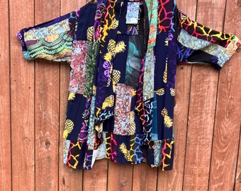 May include: A multi-colored patchwork kimono with a blue background. The kimono has a variety of patterns, including pineapples, animal prints, and floral designs. The kimono is hanging on a wooden fence.