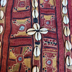 May include: Close-up of a decorative textile with a red background and geometric patterns in gold and navy blue. White seashells are sewn onto the fabric, with some arranged in a cross shape. The textile has a vintage, handcrafted appearance.