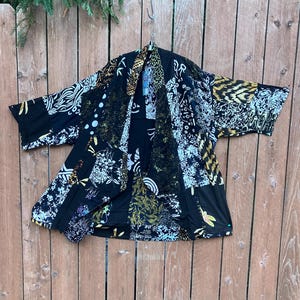 May include: A black kimono-style jacket featuring a patchwork design with various contrasting patterns in white, gold, and olive green. The garment has wide, three-quarter length sleeves, an open front, and a draped collar, showcasing a mix of floral, geometric, and abstract motifs throughout the fabric.