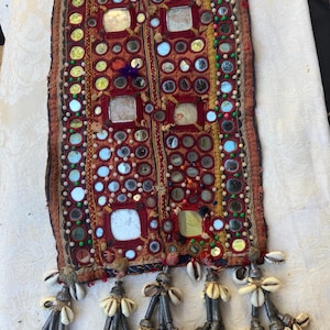 May include: A rectangular textile wall hanging featuring a rust-colored fabric base decorated with numerous small circular mirrors, square mirrors, and colorful embroidery. The bottom edge is adorned with five hanging tassels made of cowrie shells.