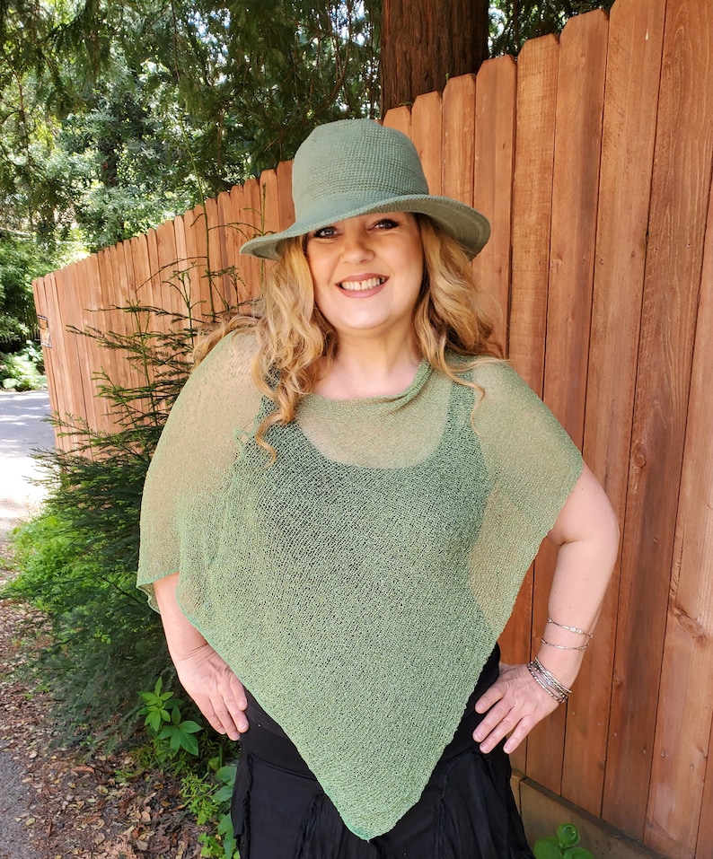 May include: A sage green knit poncho with a loose, semi-sheer texture and a pointed hemline, paired with a matching sage green bucket hat featuring a textured waffle-knit pattern and a wide, downward-sloping brim.