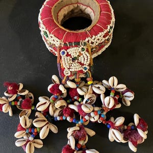 May include: A circular red fabric neck ring adorned with white seed beads and a central square beaded patch. Attached are multiple dangling strands featuring white cowrie shells, small colorful glass beads, and small red fabric pom-poms.