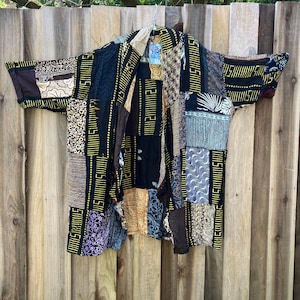 May include: A black and brown patchwork kimono with gold accents. The kimono is made of different fabrics and has a variety of patterns. It is hanging on a wooden fence.