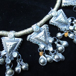 May include: A silver necklace with triangular pendants and small bells. The necklace is made of a braided cord and has a rustic, antique look.