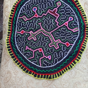 May include: A circular black fabric wall hanging with a colorful geometric design embroidered in white, green, pink, red, and blue thread. The fabric is surrounded by a green, red, and yellow border.