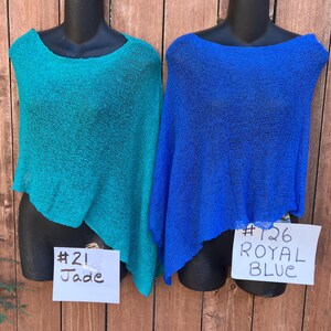 May include: Two lightweight, semi-sheer knit ponchos displayed on black mannequins. The left poncho is jade green with a tag labeled #21 Jade. The right poncho is royal blue with a tag labeled #126 ROYAL BLUE. Both garments feature a loose, open weave.