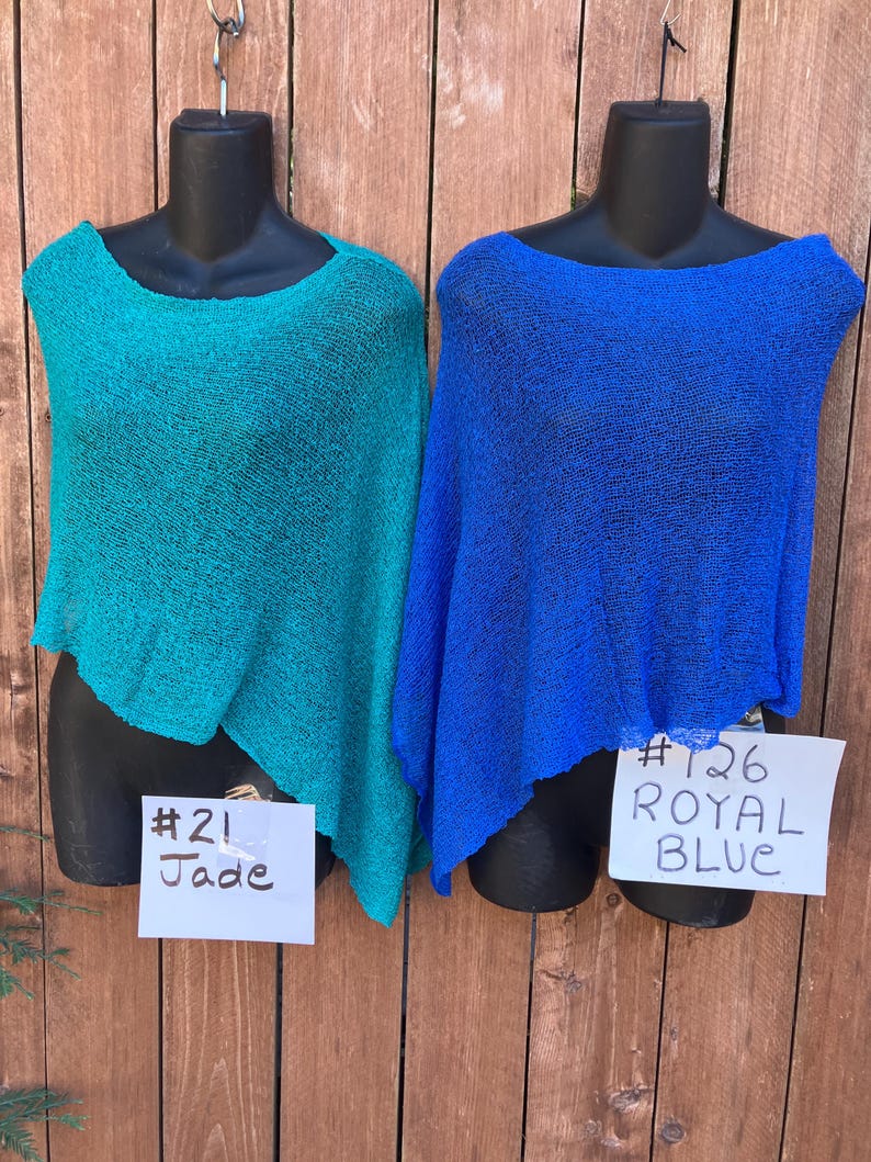 May include: Two lightweight, semi-sheer knit ponchos displayed on black mannequins. The left poncho is jade green with a tag labeled #21 Jade. The right poncho is royal blue with a tag labeled #126 ROYAL BLUE. Both garments feature a loose, open weave.