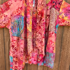 May include: A patchwork kimono-style jacket featuring a vibrant mix of pink, orange, and red fabric panels. The garment displays various intricate patterns, including floral motifs, geometric shapes, and chevron stripes, with accents of teal and yellow.
