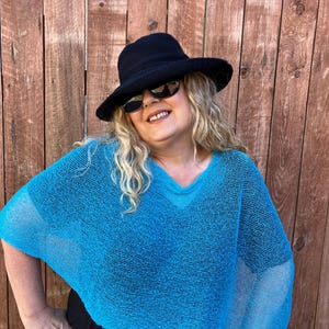 May include: A bright turquoise, loose-knit, short-sleeved poncho with a V-neckline. The garment is paired with a black wide-brimmed hat and dark-tinted sunglasses. The poncho features a semi-sheer, open-weave texture throughout the fabric.