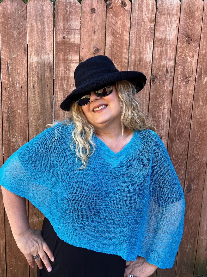 May include: A bright turquoise, loose-knit, short-sleeved poncho with a V-neckline. The garment is paired with a black wide-brimmed hat and dark-tinted sunglasses. The poncho features a semi-sheer, open-weave texture throughout the fabric.