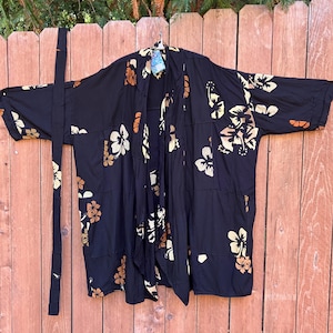 May include: Black kimono-style robe with a floral pattern in cream and brown. The robe has a tie belt and is hanging on a wooden fence. The flowers are a mix of hibiscus and other tropical blooms.