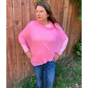 May include: A bright pink, loose-fitting, semi-sheer knit poncho with a wide neckline, worn over a light pink long-sleeved shirt and blue denim jeans. The poncho features a textured, open-weave fabric that drapes over the shoulders and torso.