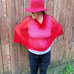 May include: A bright red crocheted bucket hat paired with a matching red, semi-sheer, loose-knit poncho featuring a cowl neckline. The poncho is worn over a white tank top and black athletic pants, creating a layered look with vibrant red textures.