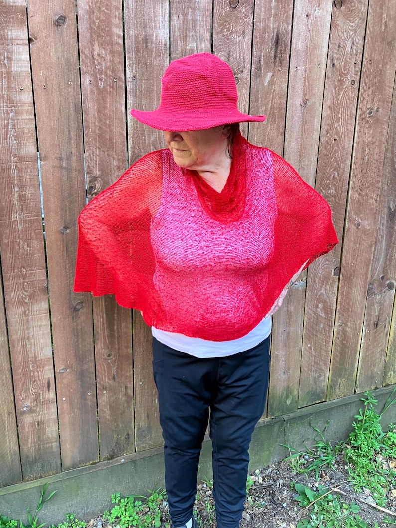 May include: A bright red crocheted bucket hat paired with a matching red, semi-sheer, loose-knit poncho featuring a cowl neckline. The poncho is worn over a white tank top and black athletic pants, creating a layered look with vibrant red textures.