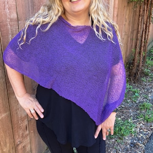 May include: A vibrant purple, loose-knit poncho featuring a wide, semi-sheer texture and a relaxed, asymmetrical drape. The garment has a simple, open neckline and short, wide sleeves that fall naturally over the shoulders, creating a lightweight layer.