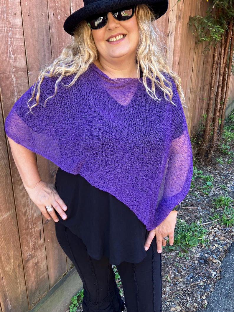 May include: A vibrant purple, loose-knit poncho featuring a wide, semi-sheer texture and a relaxed, asymmetrical drape. The garment has a simple, open neckline and short, wide sleeves that fall naturally over the shoulders, creating a lightweight layer.