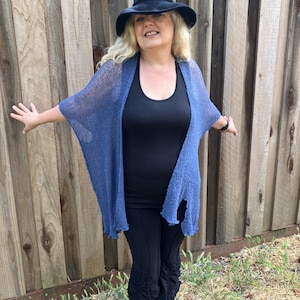 May include: A woman wearing a black tank top, black pants, and a blue shawl. She is also wearing a black hat. The background is a wooden fence and some grass.