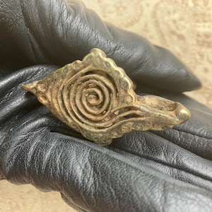 May include: A bronze-colored ring with a spiral design at the center. The ring has a decorative, ornate shape with a small, rounded end. The ring is likely made of metal and has an aged appearance.