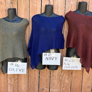 May include: Three lightweight, semi-sheer knit ponchos displayed on black mannequins. The left poncho is olive green with a sign reading #16 OLIVE. The center poncho is navy blue with a sign reading #2 NAVY. The right poncho is milk chocolate brown with a sign reading #26 Milk Chocolate.
