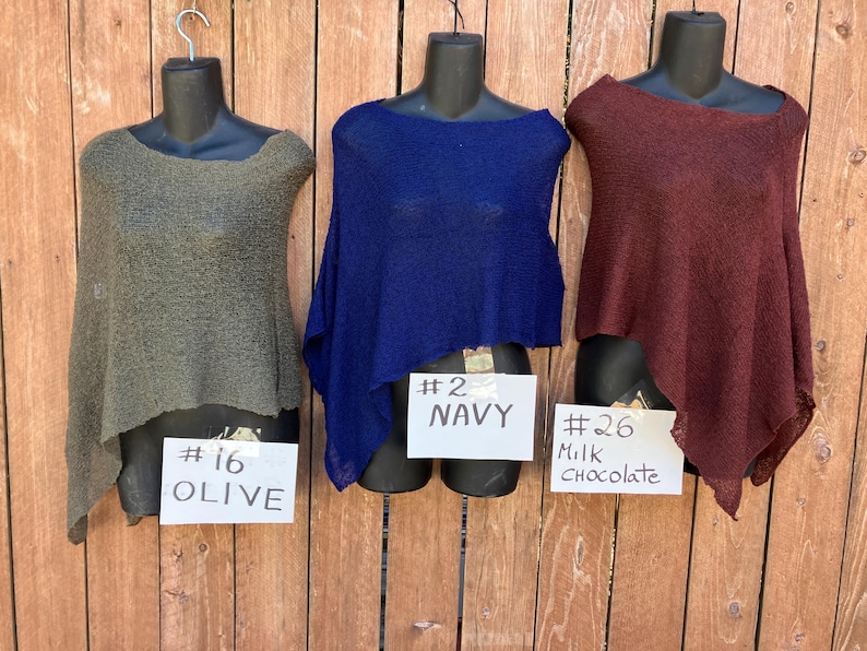 May include: Three lightweight, semi-sheer knit ponchos displayed on black mannequins. The left poncho is olive green with a sign reading #16 OLIVE. The center poncho is navy blue with a sign reading #2 NAVY. The right poncho is milk chocolate brown with a sign reading #26 Milk Chocolate.