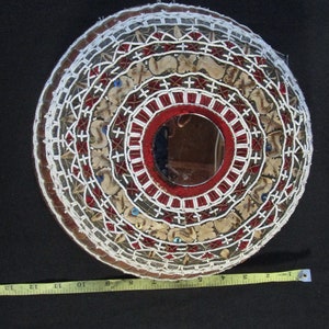 May include: A round, beaded, decorative wall hanging with a mirror in the center. The hanging is made of white beads, red beads, and brown beads. The mirror is surrounded by a red and white beaded border.