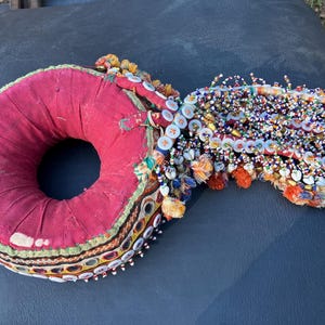 May include: A circular, red fabric cushion featuring a decorative border of small circular mirrors and white buttons. Attached to one side is a dense cluster of multi-colored glass beads, small white shells, and orange fabric tassels.