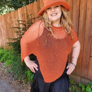 May include: A burnt orange knit hat with a wide brim and a matching burnt orange semi-sheer knit poncho featuring a loose, draped fit. The poncho is worn over a black sleeveless top and a black skirt with a layered, ruffled hemline.