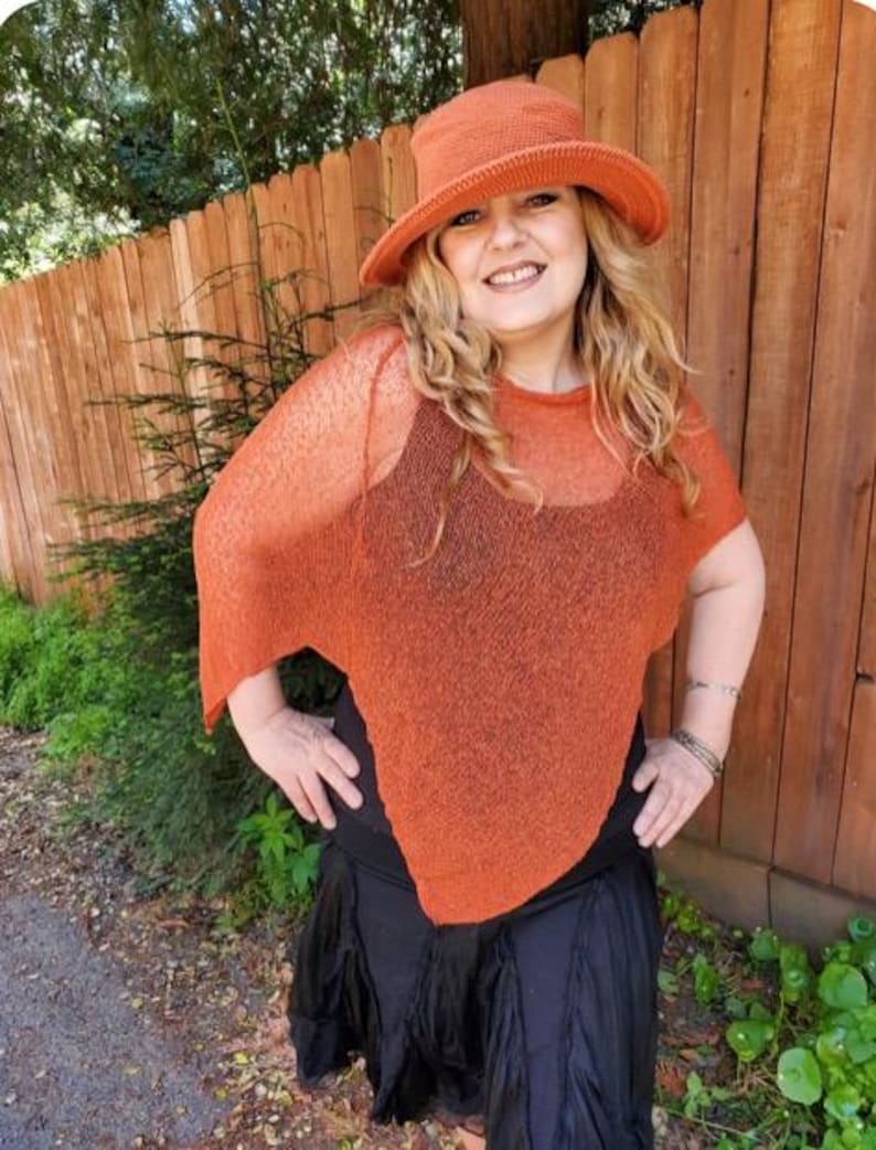 May include: A burnt orange knit hat with a wide brim and a matching burnt orange semi-sheer knit poncho featuring a loose, draped fit. The poncho is worn over a black sleeveless top and a black skirt with a layered, ruffled hemline.