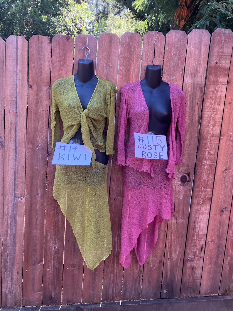 May include: Two long-sleeved, open-front knit tops and matching asymmetrical skirts displayed on mannequins. One set is kiwi green with a tag reading #17 KIWI, and the other is dusty rose with a tag reading #115 DUSTY ROSE. Both feature a textured knit.