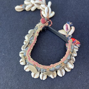 May include: An ornate, handcrafted bracelet featuring white seashells, small silver-colored metal discs, and a central band with red and gray accents. The bracelet is adorned with decorative elements and a black elastic band.