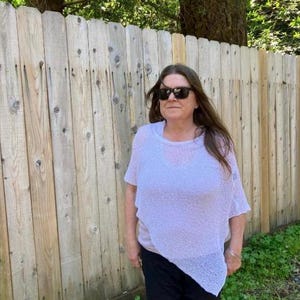 May include: A light lavender, loose-fitting, short-sleeved knit poncho with an asymmetrical hemline, worn over a solid light-colored top and dark trousers, paired with dark-tinted sunglasses.