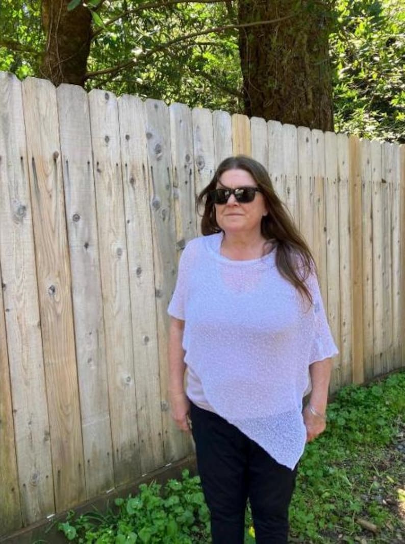 May include: A light lavender, loose-fitting, short-sleeved knit poncho with an asymmetrical hemline, worn over a solid light-colored top and dark trousers, paired with dark-tinted sunglasses.