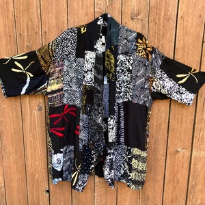 May include: A patchwork kimono-style jacket with a variety of patterns and colors. The jacket features black, white, gold, and red designs, including floral and dragonfly motifs. It hangs on a wooden background.