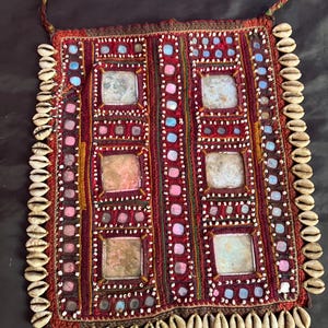 May include: A rectangular, hand-stitched textile with a deep red background. The design features square mirrored accents, surrounded by pink, blue, and white beads. The edges are adorned with numerous white seashells, and the piece has a woven loop at the top.
