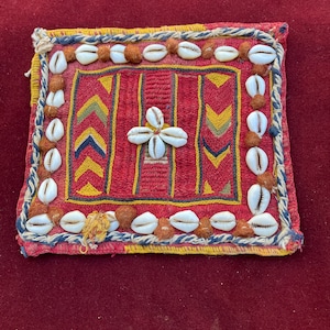 May include: A red, yellow, and blue embroidered fabric square with a white shell design in the center. The fabric is decorated with a border of white shells.