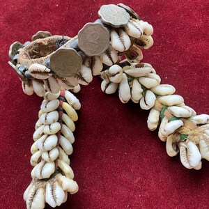 May include: An ornate bracelet featuring white seashells and silver coins. The bracelet is constructed with a woven fabric base and adorned with multiple strands of seashells. The coins are attached to the bracelet's surface, adding a unique decorative element.