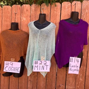 May include: Three knit ponchos displayed on black mannequins. The left poncho is cognac brown with a tag labeled #9 COGNAC. The center poncho is mint green with a tag labeled #22 MINT. The right poncho is purple with a tag labeled #14 PURPLE ROYALE.