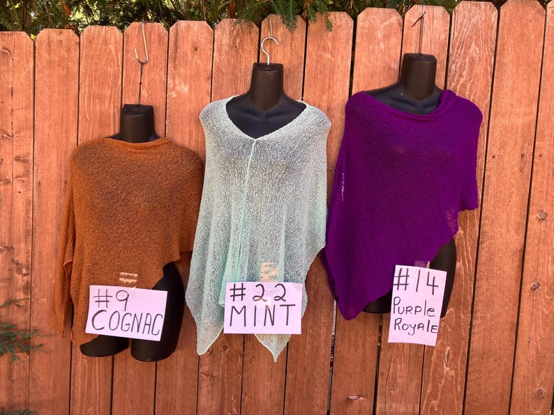 May include: Three knit ponchos displayed on black mannequins. The left poncho is cognac brown with a tag labeled #9 COGNAC. The center poncho is mint green with a tag labeled #22 MINT. The right poncho is purple with a tag labeled #14 PURPLE ROYALE.