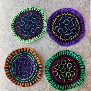 May include: Four circular fabric coasters with black centers and embroidered patterns. Each coaster features a distinct neon-colored stitched border and contrasting thread designs in blue, orange, purple, yellow, and red, creating a vibrant set.