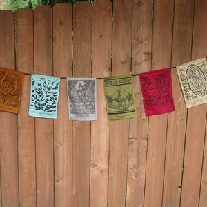 May include: Seven colorful fabric prayer flags hanging on a wooden fence. The flags feature different designs and text, including "serenity", "compassion", "peace", and "much".