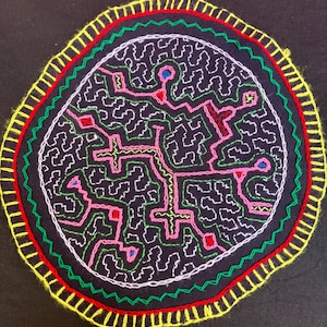 May include: A circular black fabric piece featuring intricate hand-stitched embroidery in white, pink, and green. The border consists of concentric rings in red and yellow. The central design displays a complex, maze-like pattern of lines.