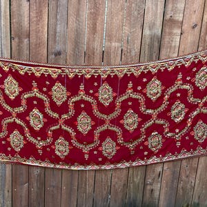 May include: A rectangular, red textile with intricate gold embroidery. The design features repeating floral and geometric patterns. The edges are trimmed with gold beading. The textile is displayed against a wooden background.