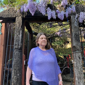May include: A light purple, semi-sheer, knitted poncho with an asymmetrical hemline. The garment features a loose, draped fit and a fine, textured knit pattern. It is worn over a light-colored top and dark trousers, showcasing a lightweight design.