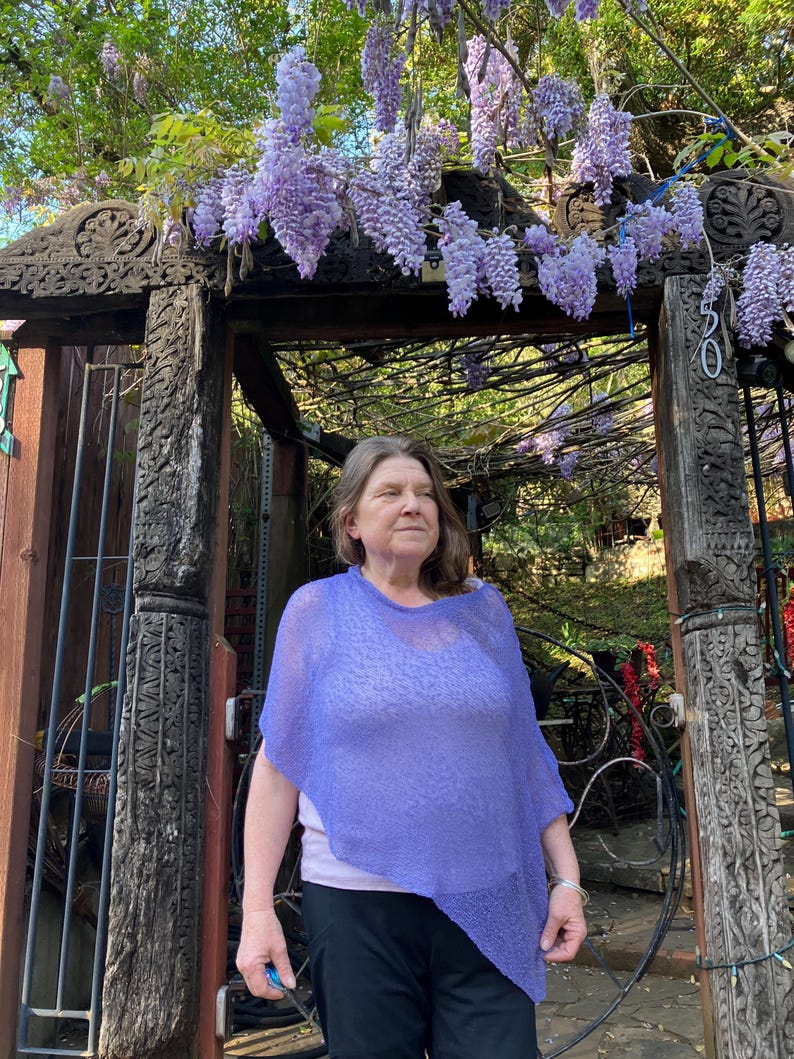 May include: A light purple, semi-sheer, knitted poncho with an asymmetrical hemline. The garment features a loose, draped fit and a fine, textured knit pattern. It is worn over a light-colored top and dark trousers, showcasing a lightweight design.