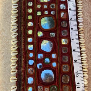 May include: A rectangular dark red fabric wall hanging decorated with numerous small inset mirrors and bordered by white cowrie shells. A white measuring tape placed along the side indicates a length of approximately 13.5 inches.