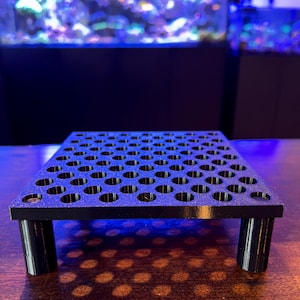 Puede incluir: Un soporte frag negro y rectangular para acuarios, con numerosos agujeros circulares. Se apoya en cuatro patas cilíndricas. El fondo muestra un acuario con peces y corales coloridos.