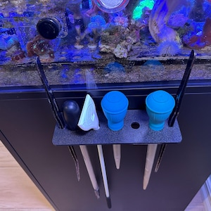 Könnte beinhalten: Ein schwarzer Aquarienstand mit einem schwarzen Regal, das verschiedene Aquarienwerkzeuge enthält. Die Werkzeuge umfassen einen schwarzen Schaber, einen weißen Schaber, zwei blaue Scheuerbürsten und drei lange Reinigungswerkzeuge. Das Aquarium ist oben sichtbar.